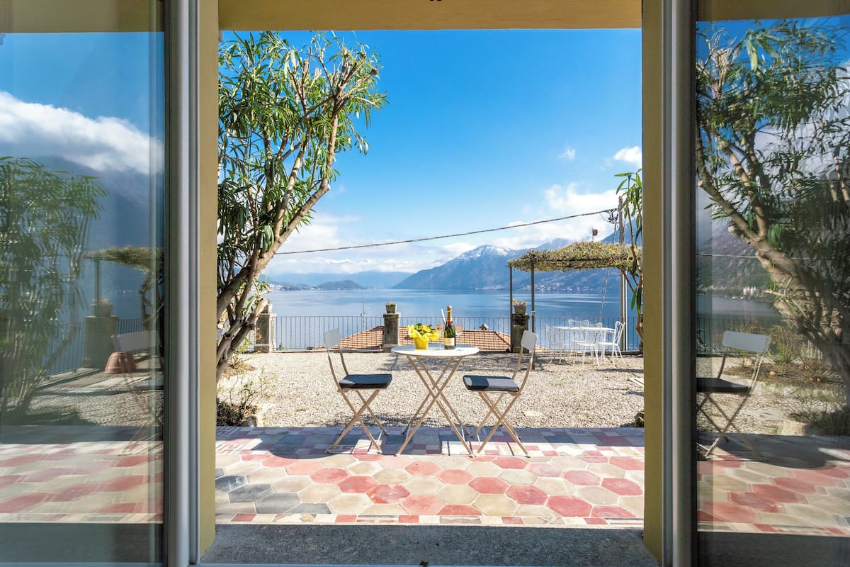 A patio with a table and chairs, a bottle of wine, and a view of a lake and mountains.