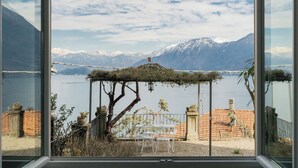 Vista a través de una ventana que muestra una terraza con mesa y sillas, una pérgola con plantas y montañas al fondo.