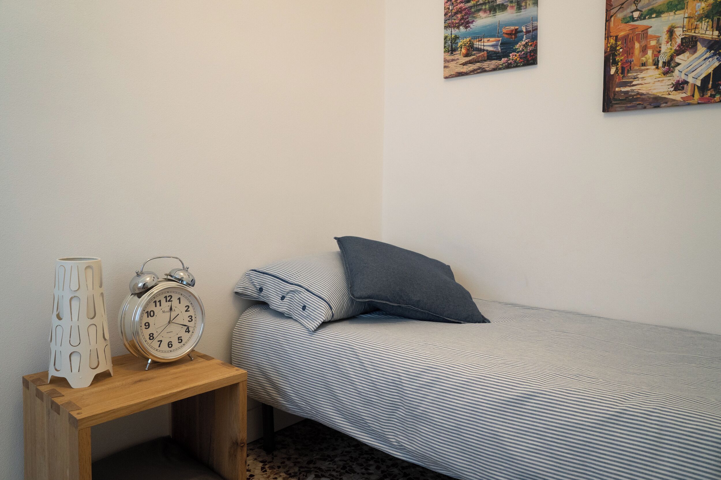 Une petite chambre à coucher, bien rangée, avec un lit simple, une table de chevet sur laquelle se trouvent une horloge et un vase décoratif, ainsi que deux tableaux accrochés au mur.