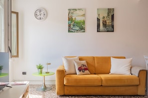 Un salon avec un canapé jaune, une petite table ronde ornée d’une plante et deux tableaux accrochés au mur.