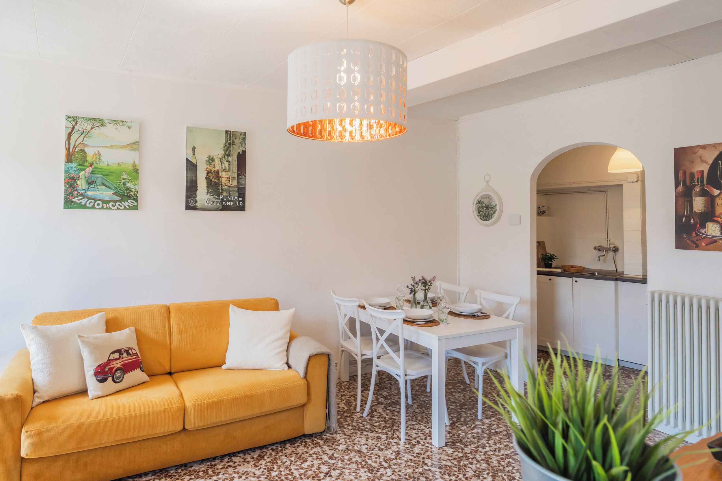 Una sala de estar con un sofá amarillo, una mesa de comedor blanca y sillas, una lámpara de techo blanca y dos cuadros colgados en la pared.