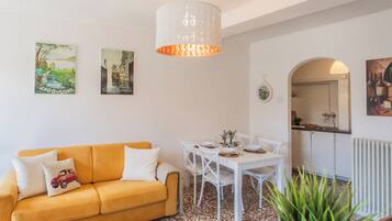 Una sala de estar con un sofá amarillo, una mesa de comedor blanca y sillas, una lámpara de techo blanca y dos cuadros colgados en la pared.