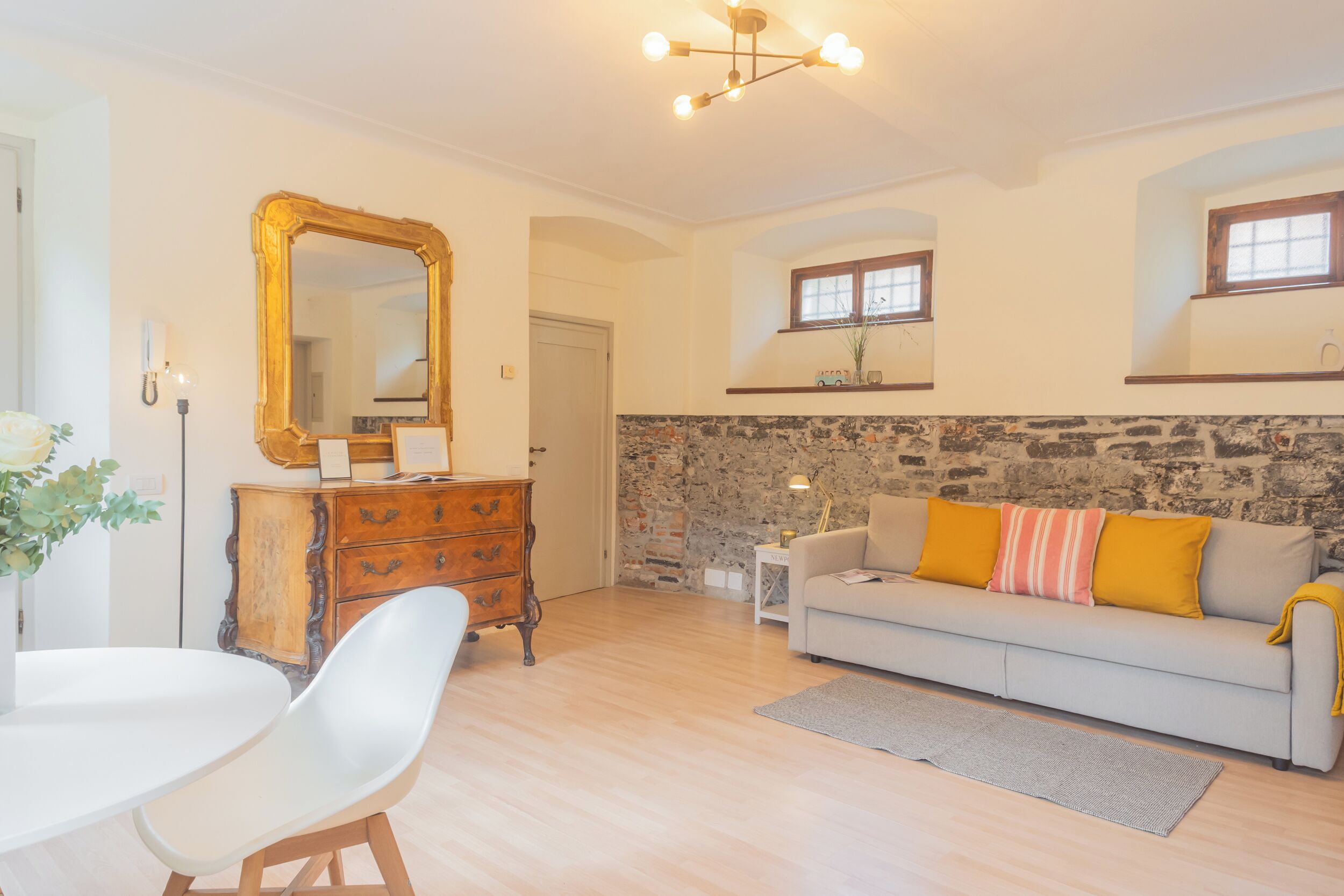 Una sala de estar con una pared de piedra, un sofá con cojines amarillos, una mesa blanca con sillas y un espejo grande.