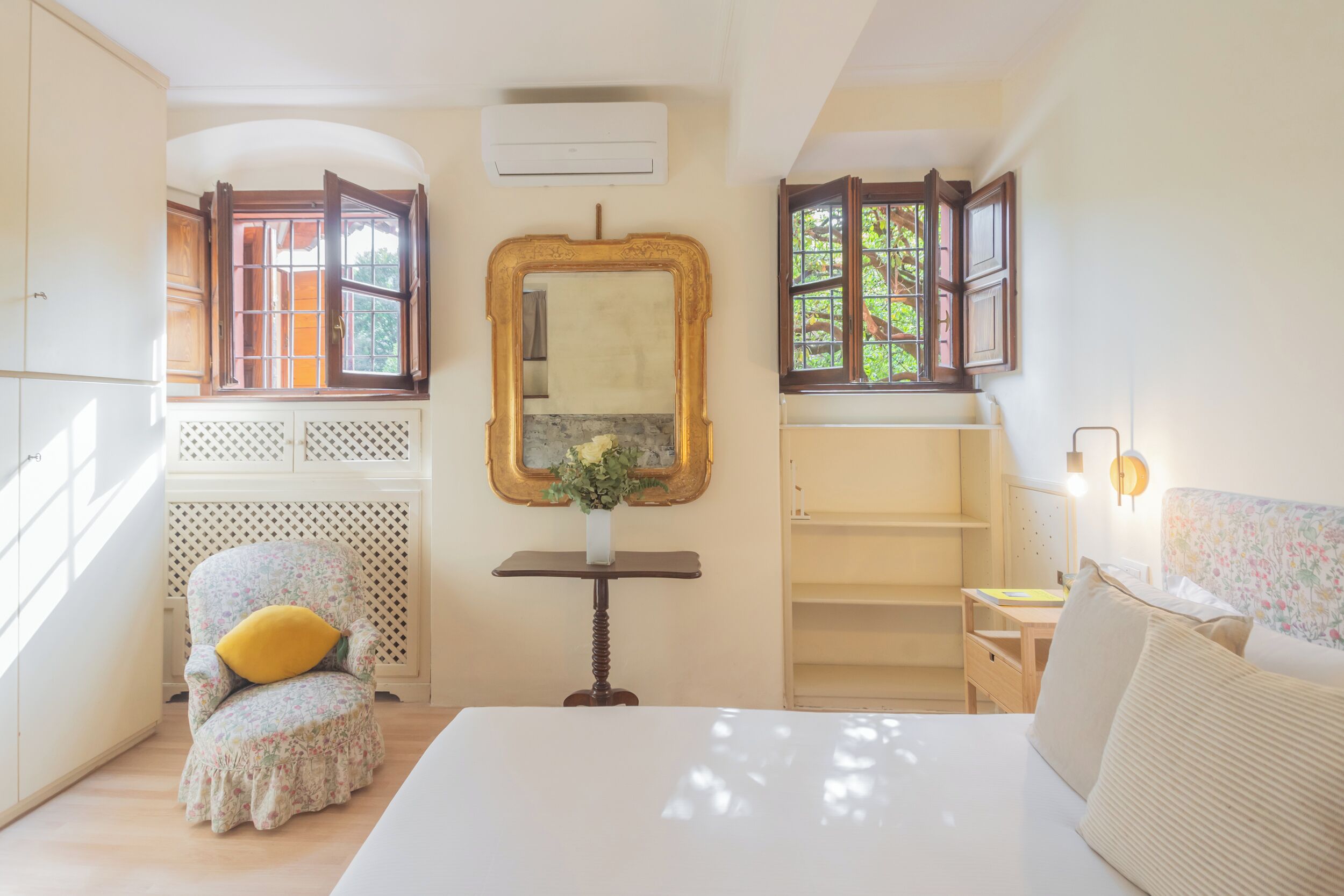 Un dormitorio con cama, una silla con cojín, una mesa con un florero y un espejo.