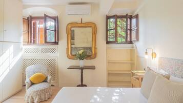 Un dormitorio con cama, una silla con cojín, una mesa con un florero y un espejo.