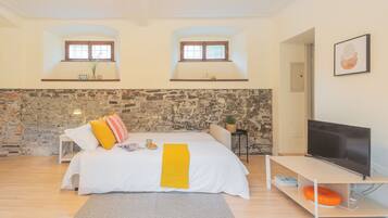 Un dormitorio con pared de piedra, una cama con ropa de cama blanca y almohadas coloridas, un televisor sobre un mueble bajo y una mesita con una planta.