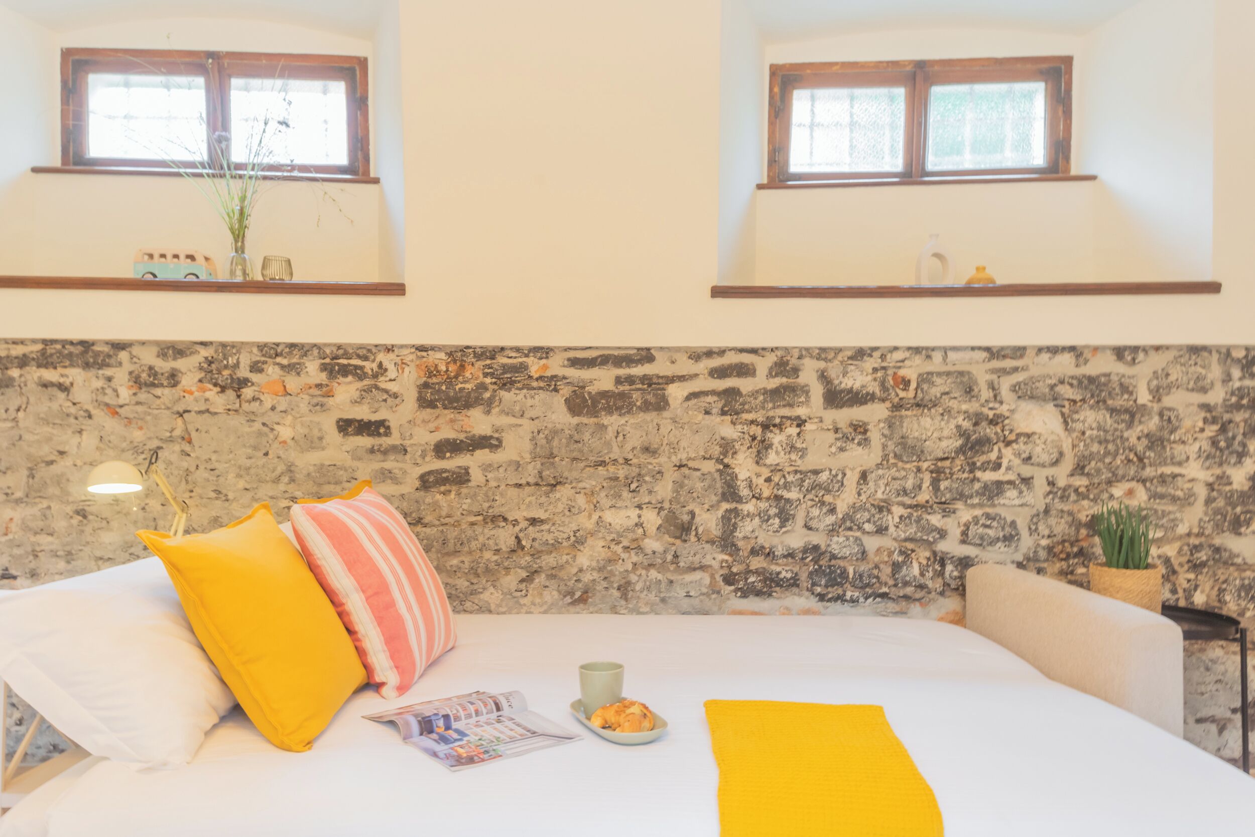 Un dormitorio con pared de piedra, cama con ropa de cama blanca, almohadas amarillas y una mesita con una taza y un plato.