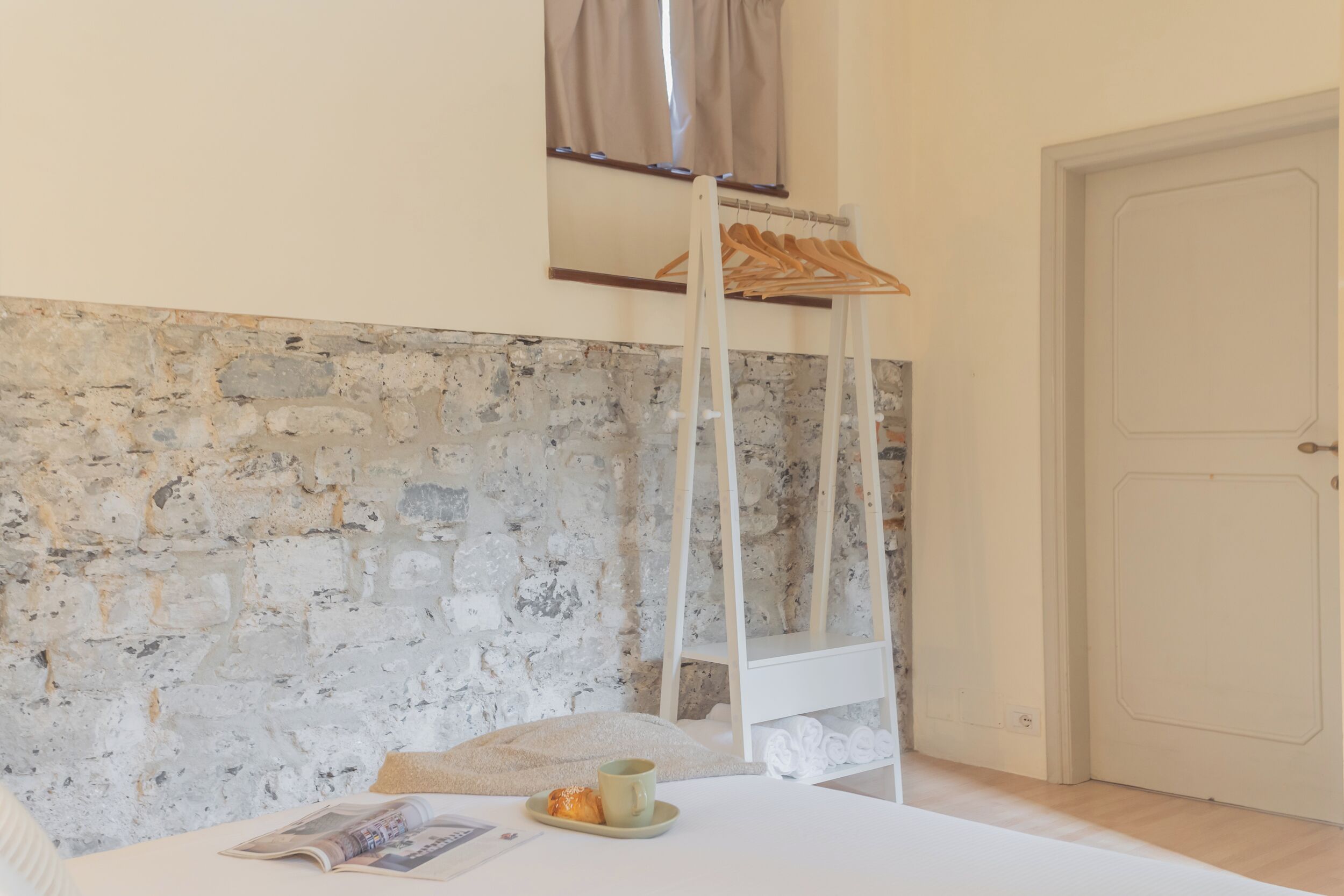 A room with a stone wall, a bed with a pillow and a cup, a wooden hanger, and a door.