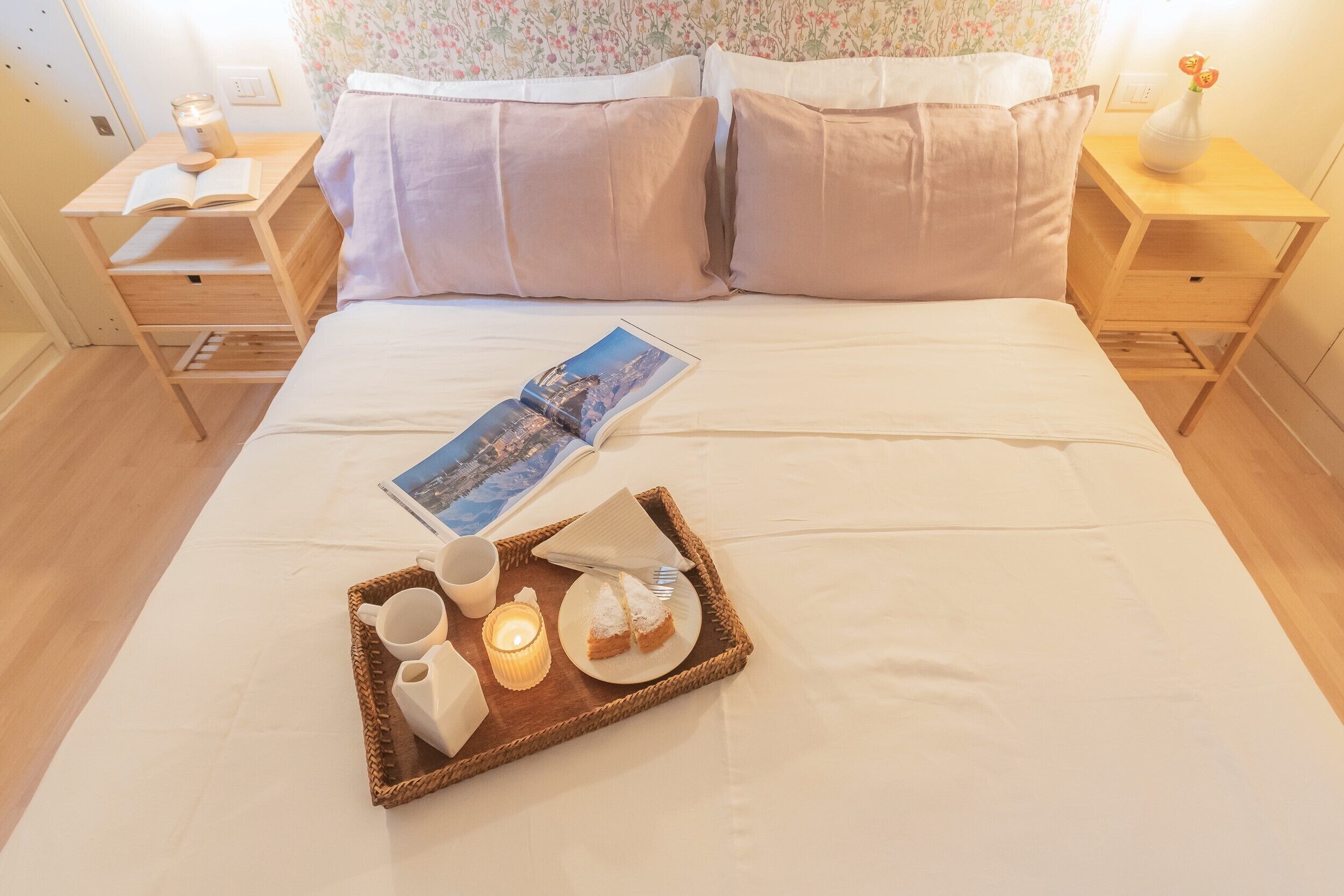 Una cama bien hecha con una bandeja de desayuno, un libro y una vela sobre una mesita de noche.