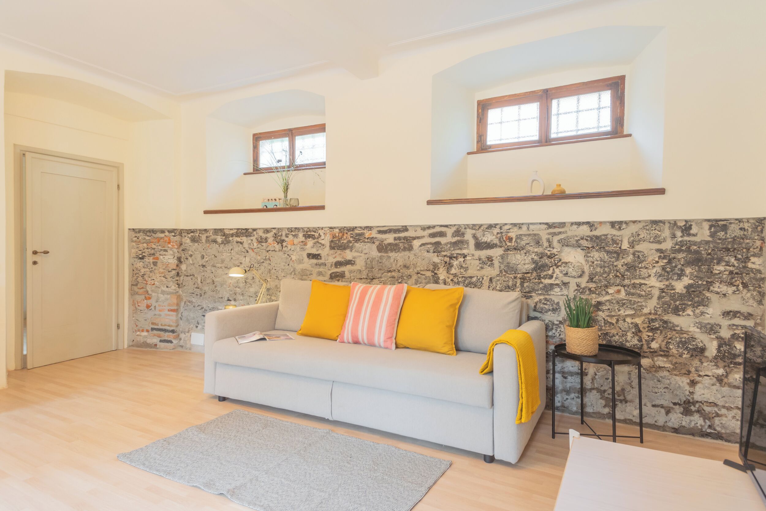 A living room with a stone wall, a sofa with yellow cushions, a small table with a plant, and a door.