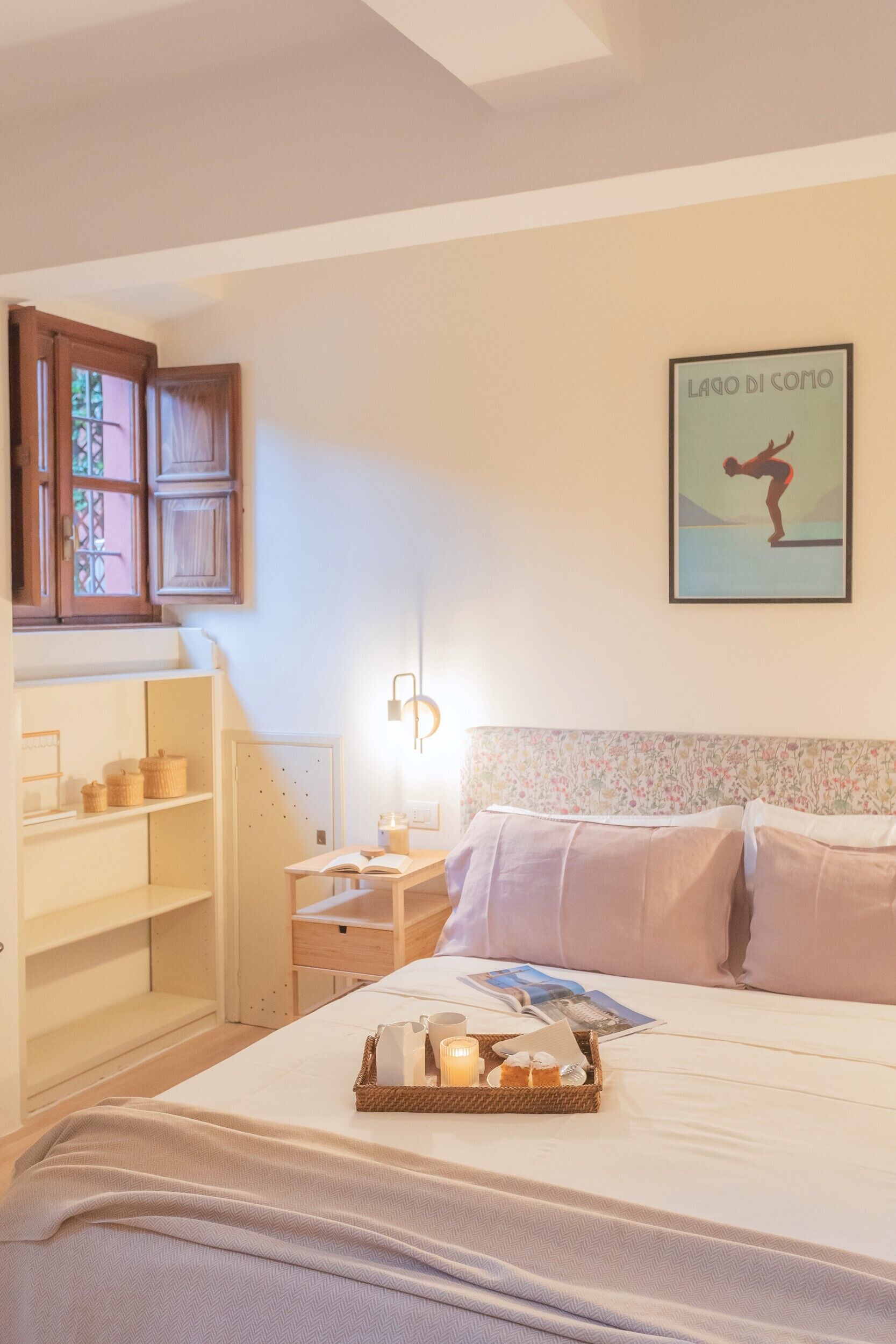 A bedroom with a bed, a nightstand, a lamp, a window with curtains, a framed picture on the wall, and a tray with candles and a book.