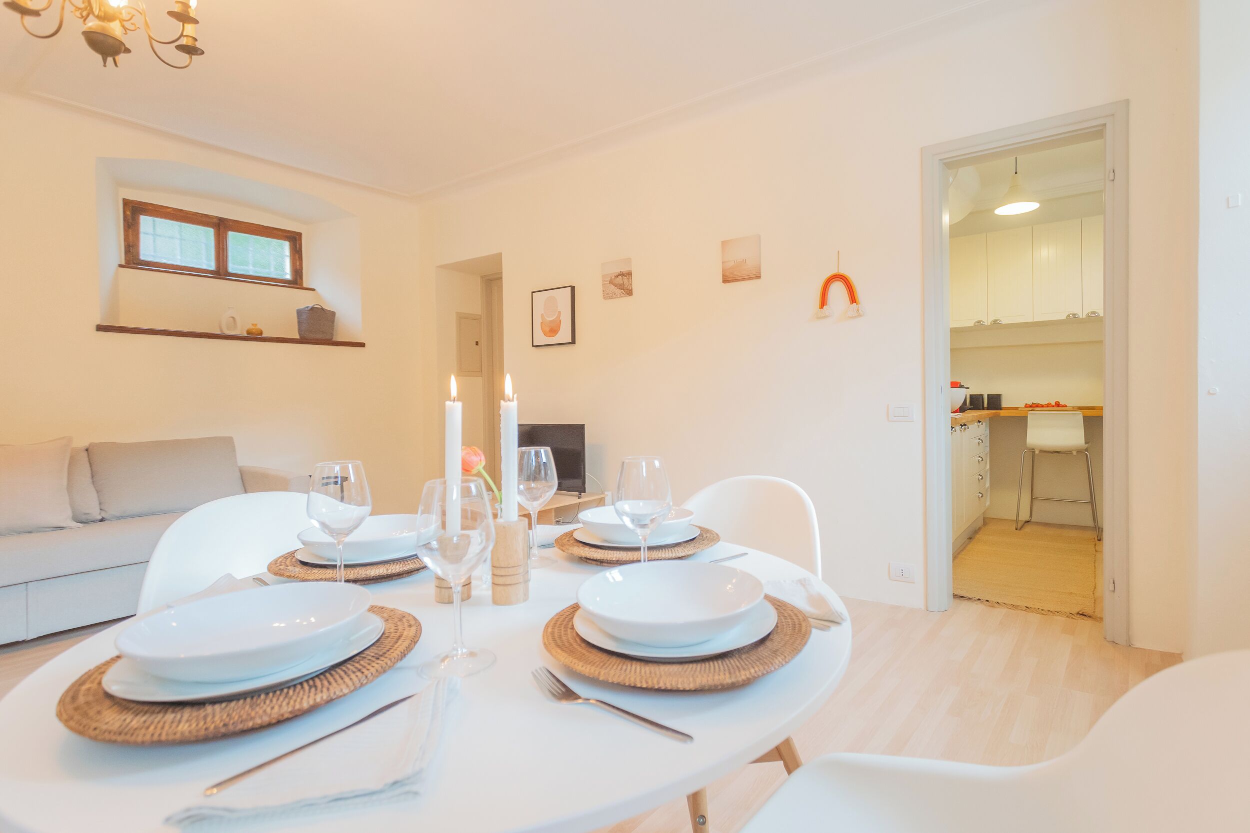 A dining area with a table set for four, white plates, and wine glasses. There is a sofa and a kitchen visible in the background.