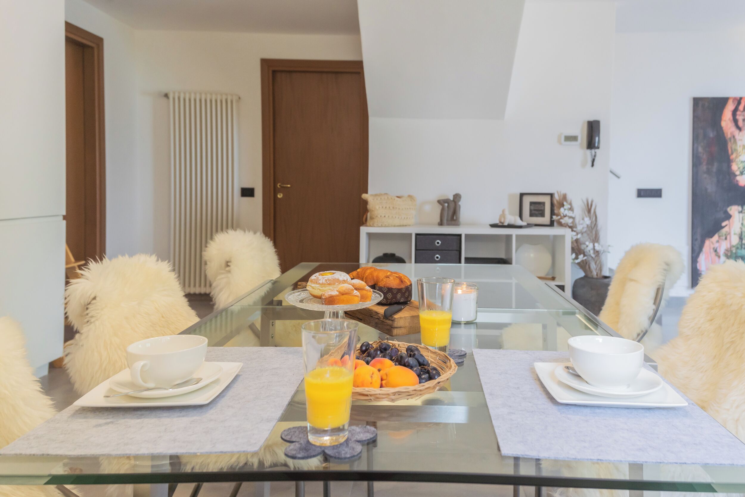 Een eetruimte met een glazen tafel, witte stoelen, en een ontbijt dat croissants, gebak en een fruitmand bevat.