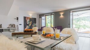 A modern dining area with a glass table, white fluffy chairs, a painting on the wall, and a view of greenery outside. - Harmony on the Lake by Rent all Como (Argegno)