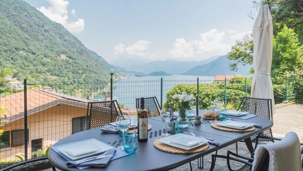 A table set outdoors with a view of a lake and mountains, surrounded by a fence. - Harmony on the Lake by Rent all Como (Argegno)
