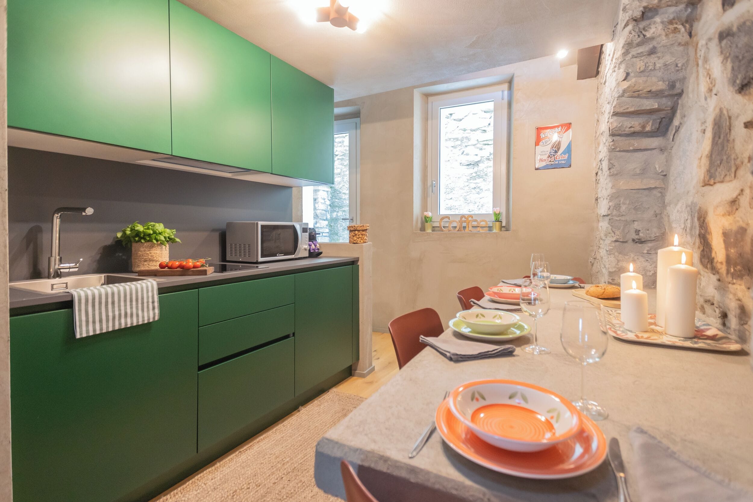 A modern kitchen with green cabinets, a microwave, and a dining area with a table set for two, candles, and a window with a view of trees.