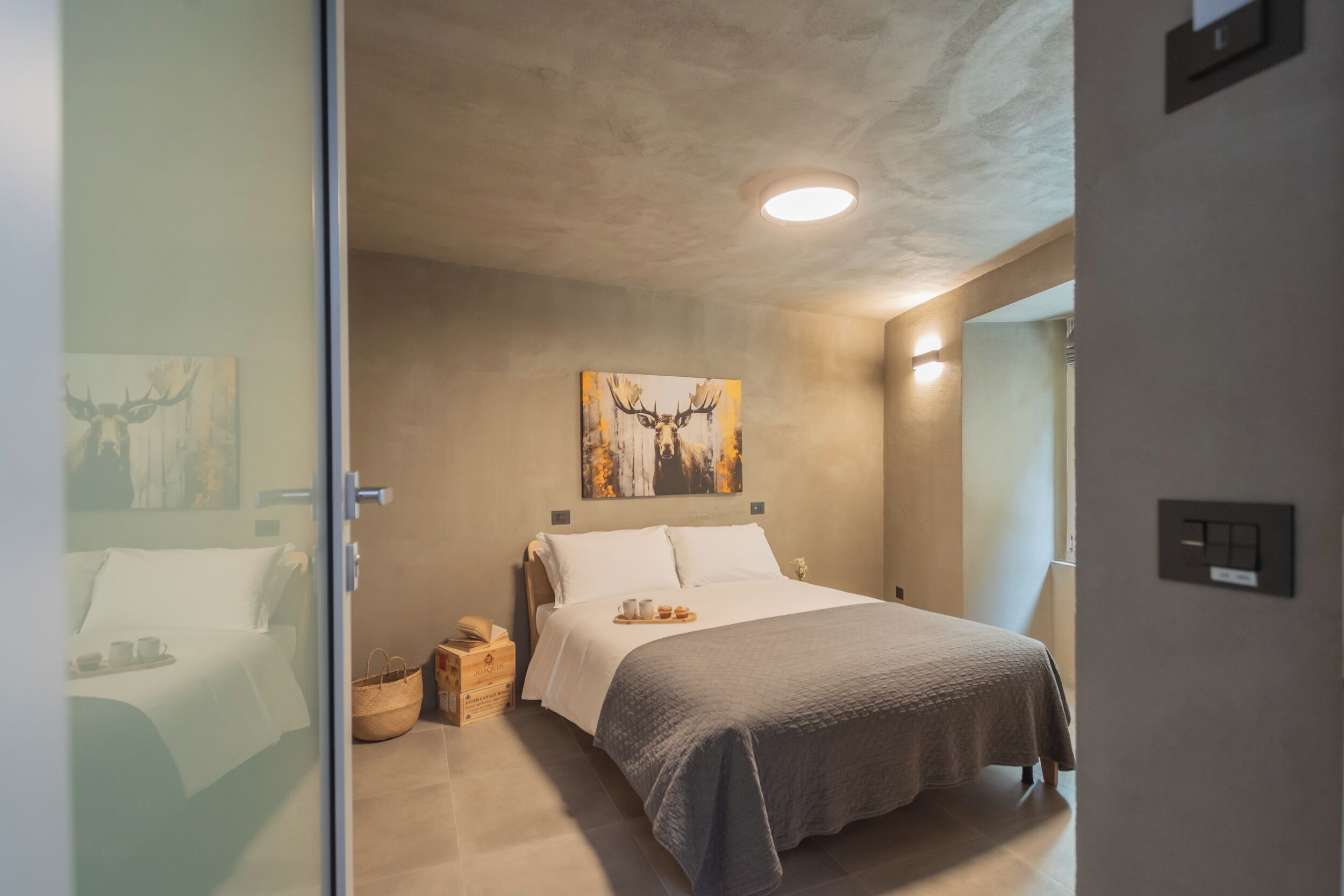 A modern bedroom with a large bed, a painting of a deer, a bedside table with a lamp, and a window with curtains.