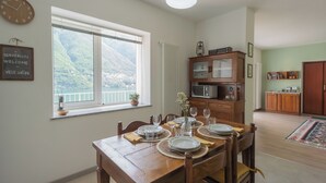 A dining area with a wooden table set for four, a view of mountains through a large window, and a kitchen in the background. - La Quiete With Private Garden by Rent all Como (Laglio)