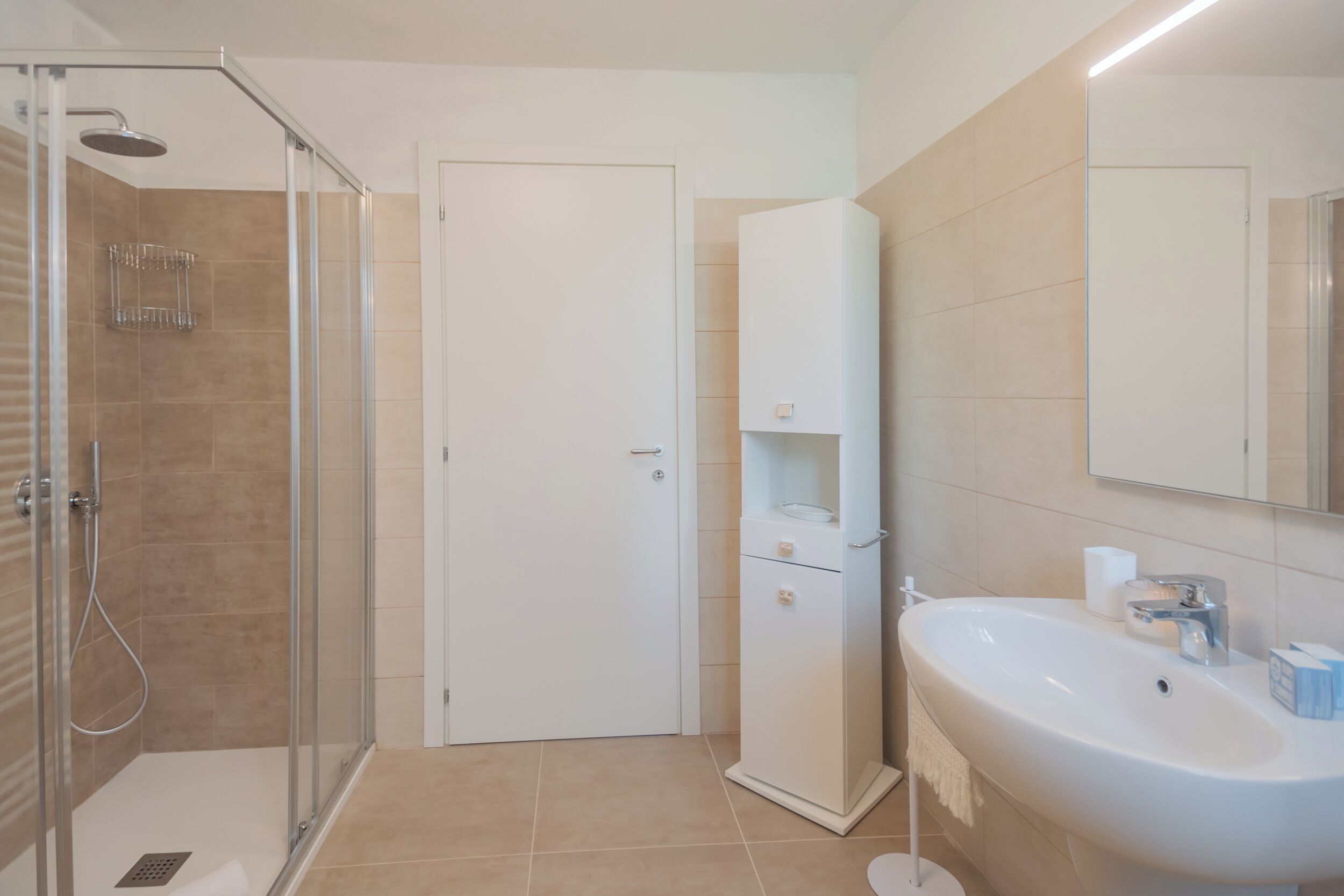 A modern bathroom with a glass-enclosed shower, a white sink with a mirror above, and a cabinet with a towel hanging on the door.