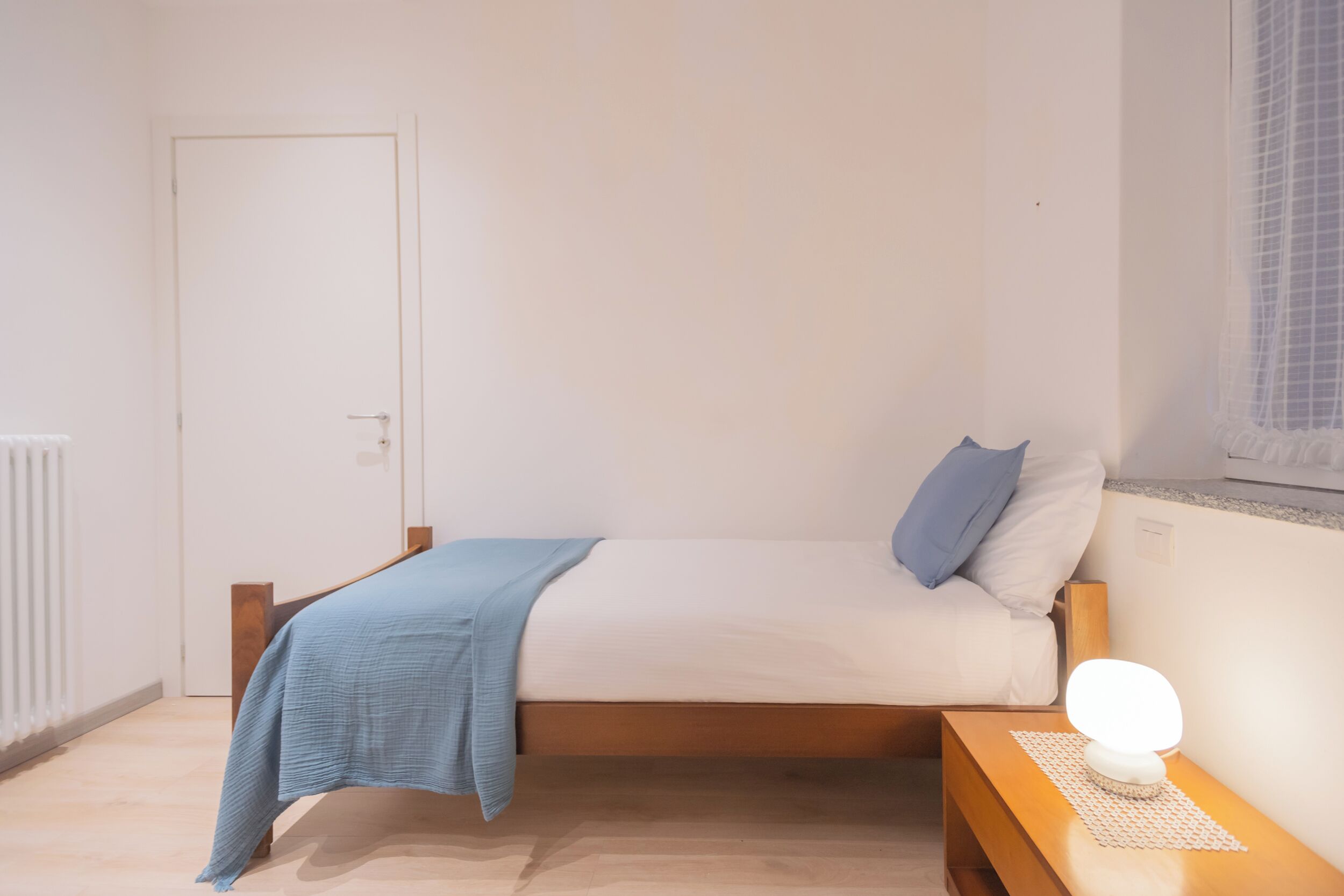 A minimalist bedroom with a single bed, a bedside table with a lamp, and a window with a view.