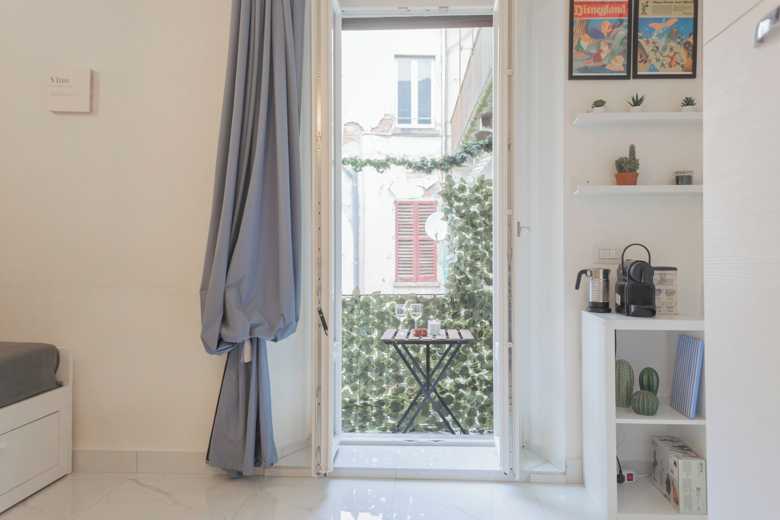 Una habitación con una cama, una mesita con un vaso, un estante con objetos decorativos y vistas a un edificio con un persiano rojo.