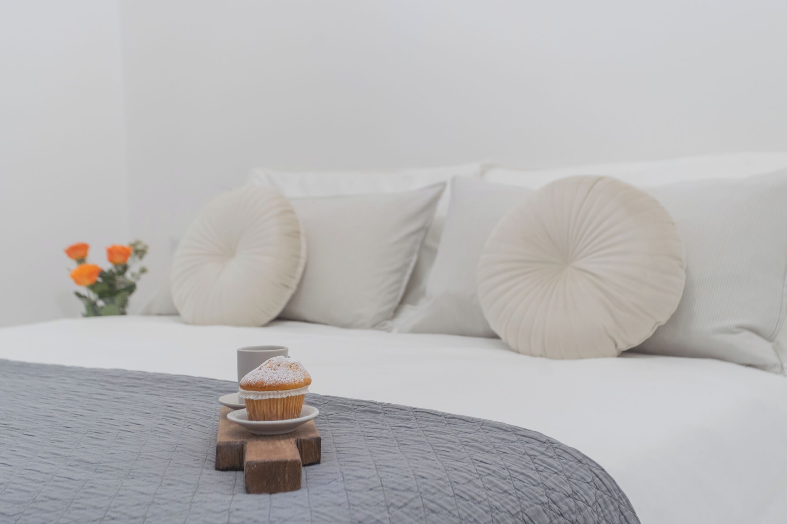 A neatly made bed with a grey quilt, a wooden stand holding a cupcake, and a vase with orange flowers.