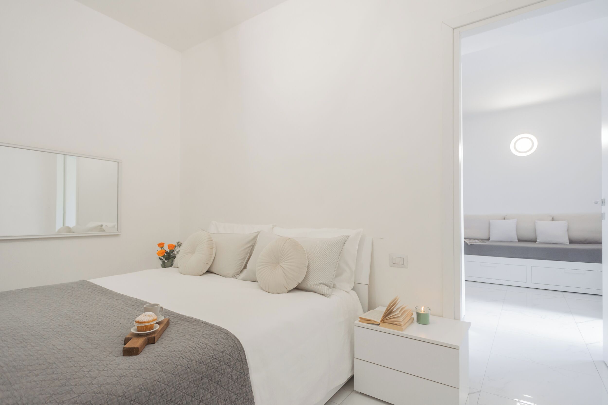 Une chambre moderne avec un lit soigneusement fait, une table de chevet sur laquelle se trouvent un livre et une bougie, et une vue sur un espace de vie adjacent.