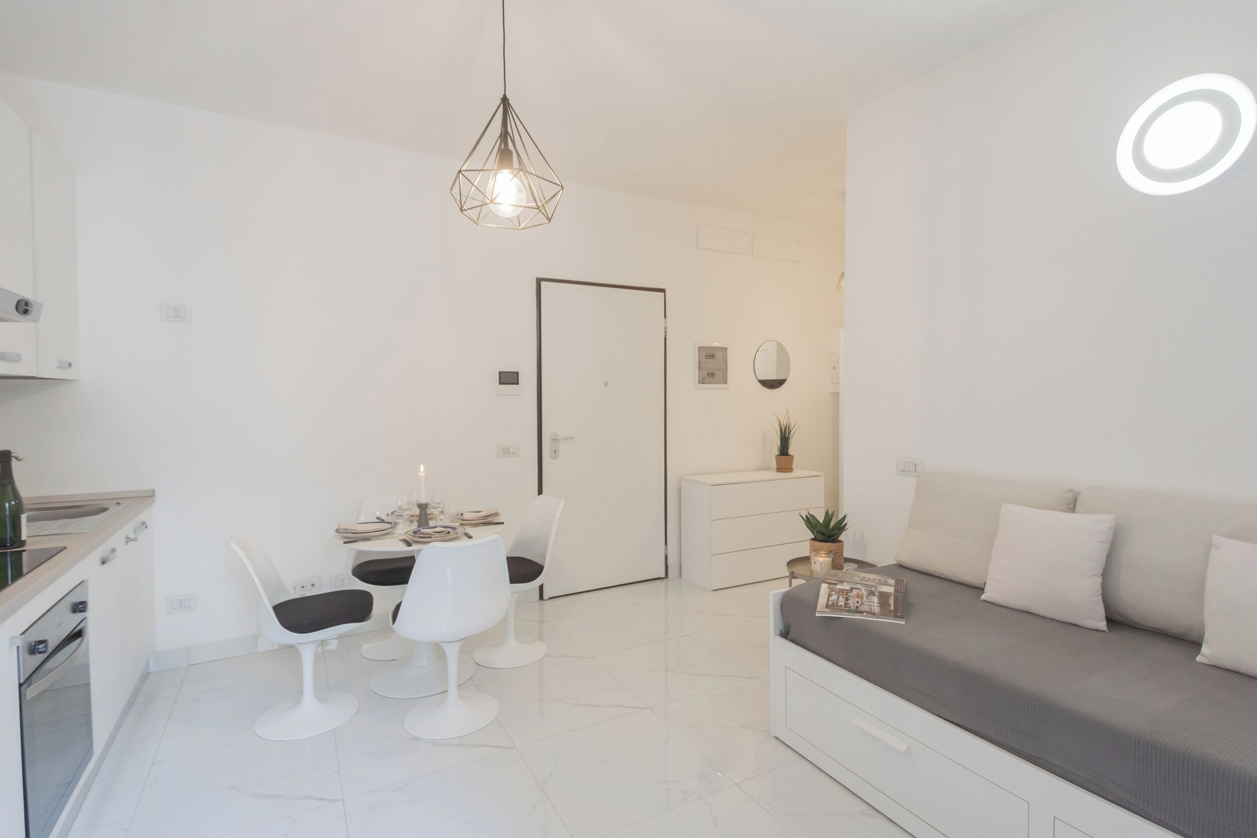 A modern, compact living space with a kitchenette, a dining area with white chairs, a grey sofa, and a pendant light.