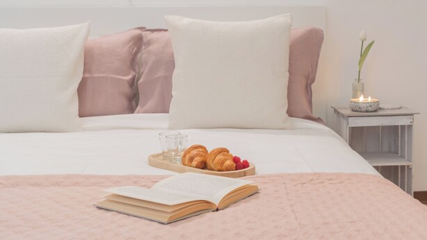 A neatly made bed with a pink blanket, two pillows, a tray with croissants and raspberries, a lit candle, and an open book on the bed. - Nonna Tere Apartment by Rent all Como (Carate Urio)