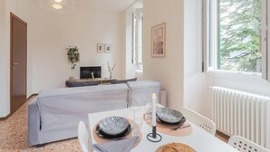 A modern hotel room with a dining area, a bed, a TV, and a window with a view of trees. - Nonna Tere Apartment by Rent all Como (Carate Urio)