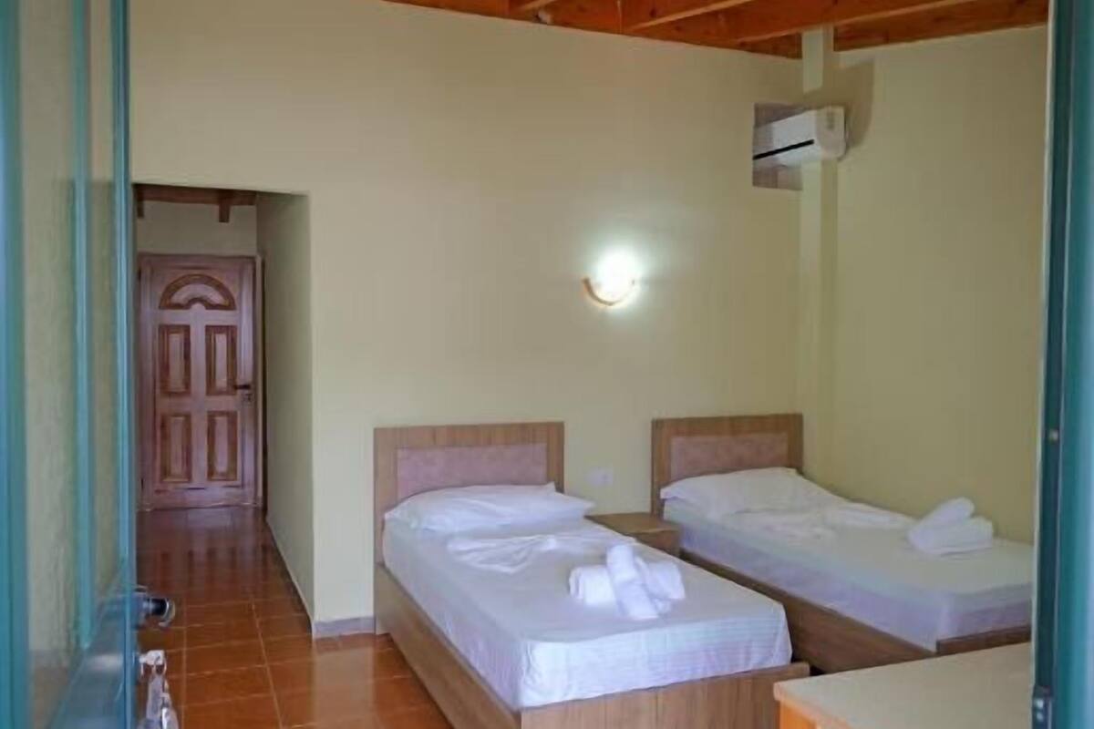 A room with two beds, a wooden ceiling, a tiled floor, and a small kitchenette.