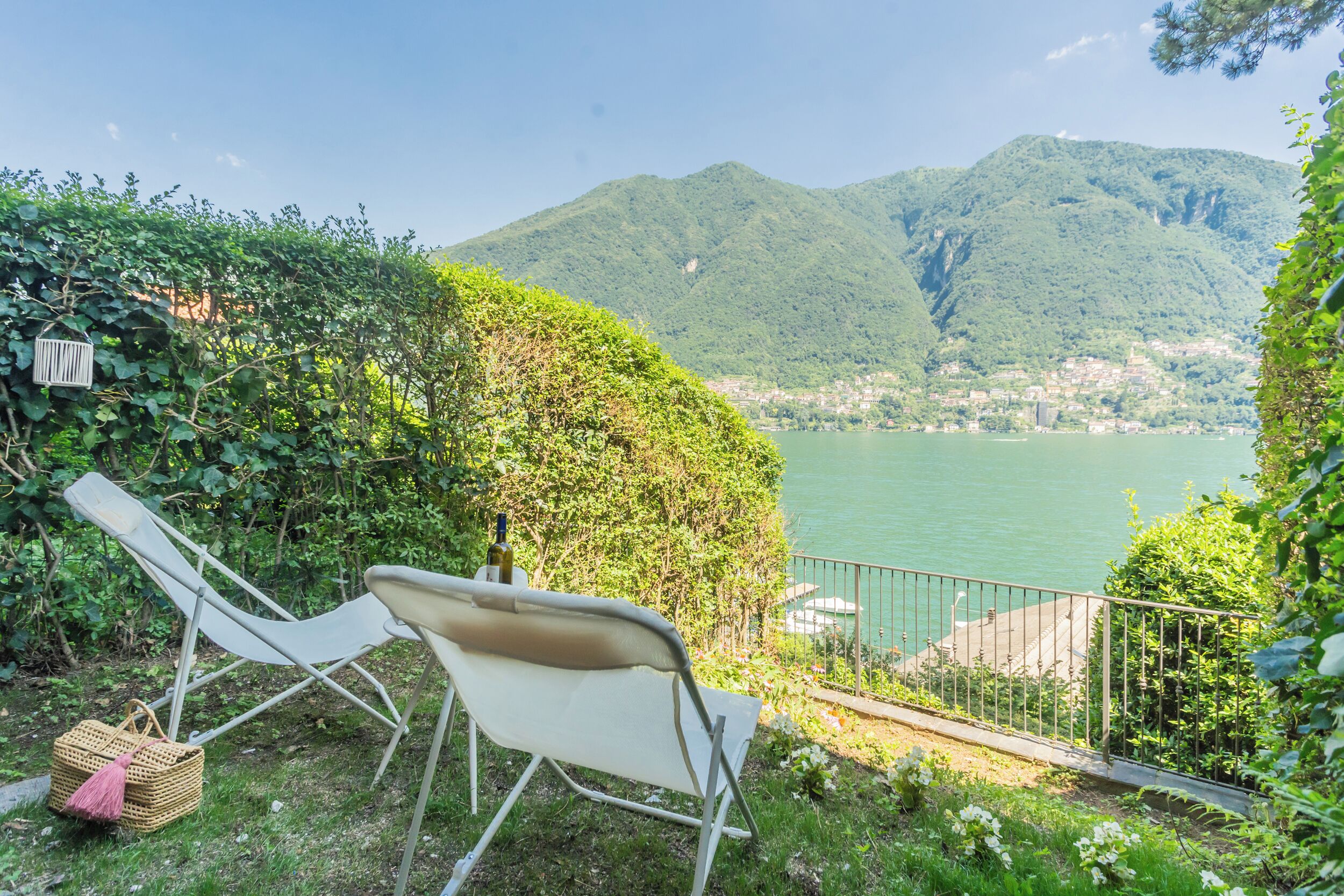 Een balkon met uitzicht op een meer en bergen, twee lounge fauteuils en een fles wijn.