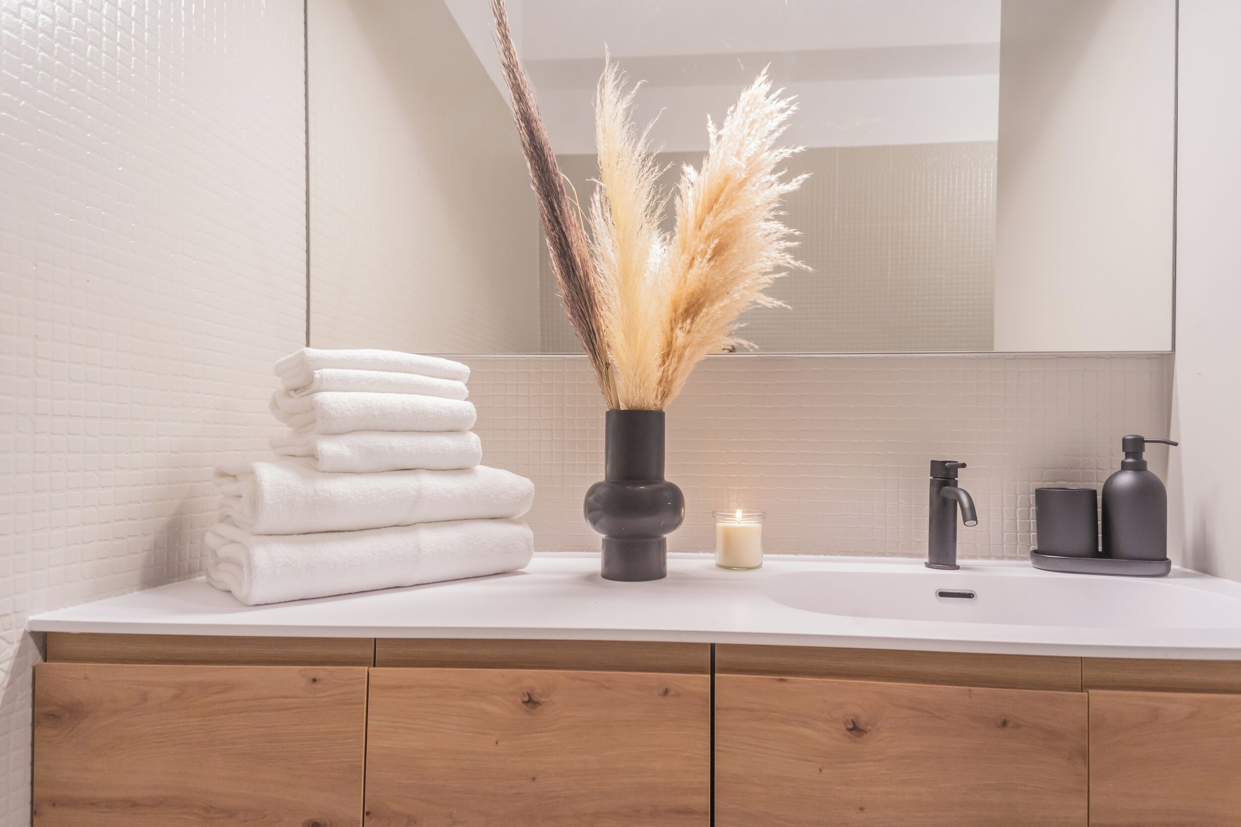 Un bagno con un lavandino bianco, uno specchio, una pila di asciugamani bianchi, una candela e un vaso con pampa.