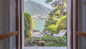 Vista desde una habitación con puerta de vidrio, que da a un área junto al lago con dos sillas y una mesa, una botella y una cesta.