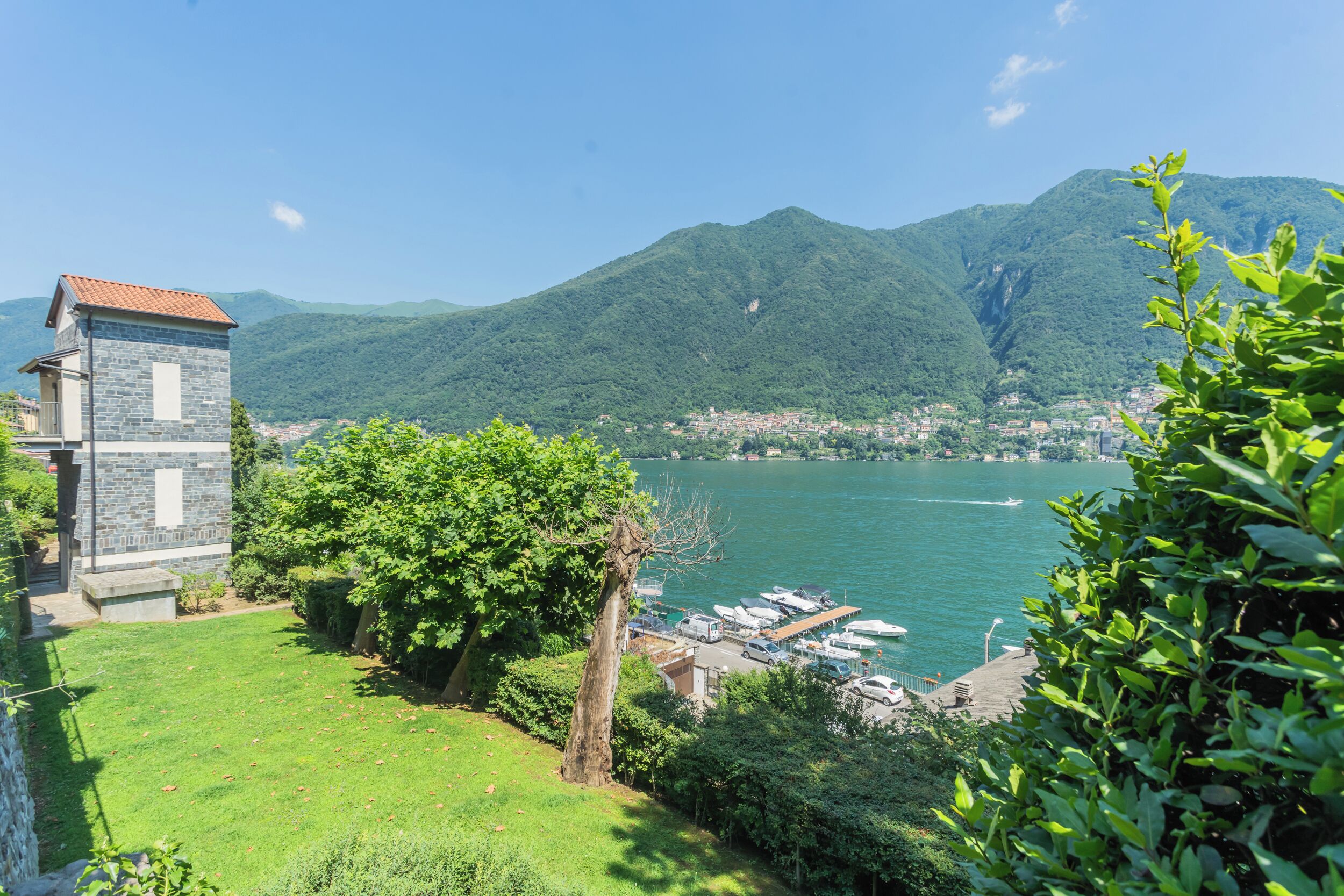 A lakeside view with a stone building, lush greenery, and boats docked near the shore.