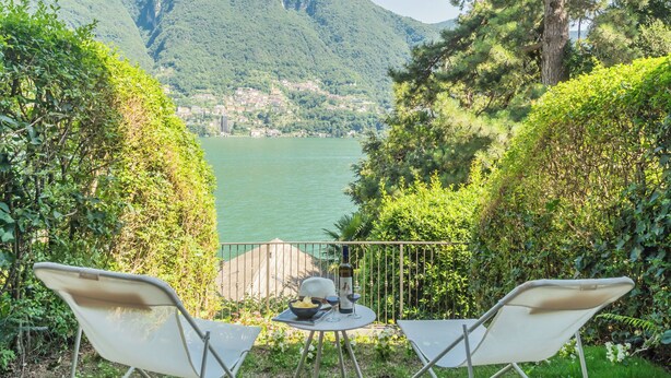 Una terraza con dos sillas, una mesa y una botella de vino, con vistas a un lago y montañas al fondo.