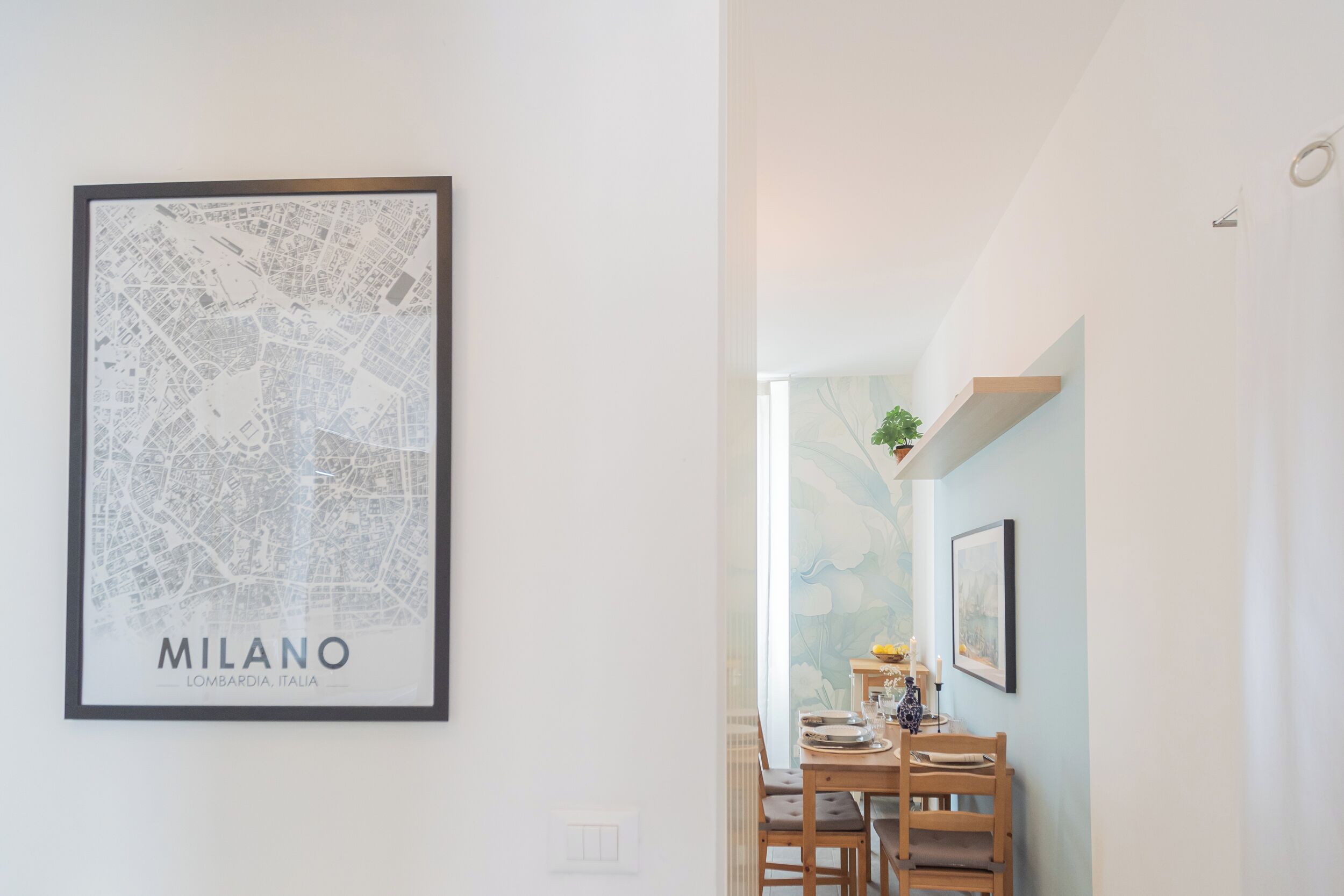 A framed city map of Milano on a white wall, a dining area with a wooden table and chairs, and a wall with a floral mural.