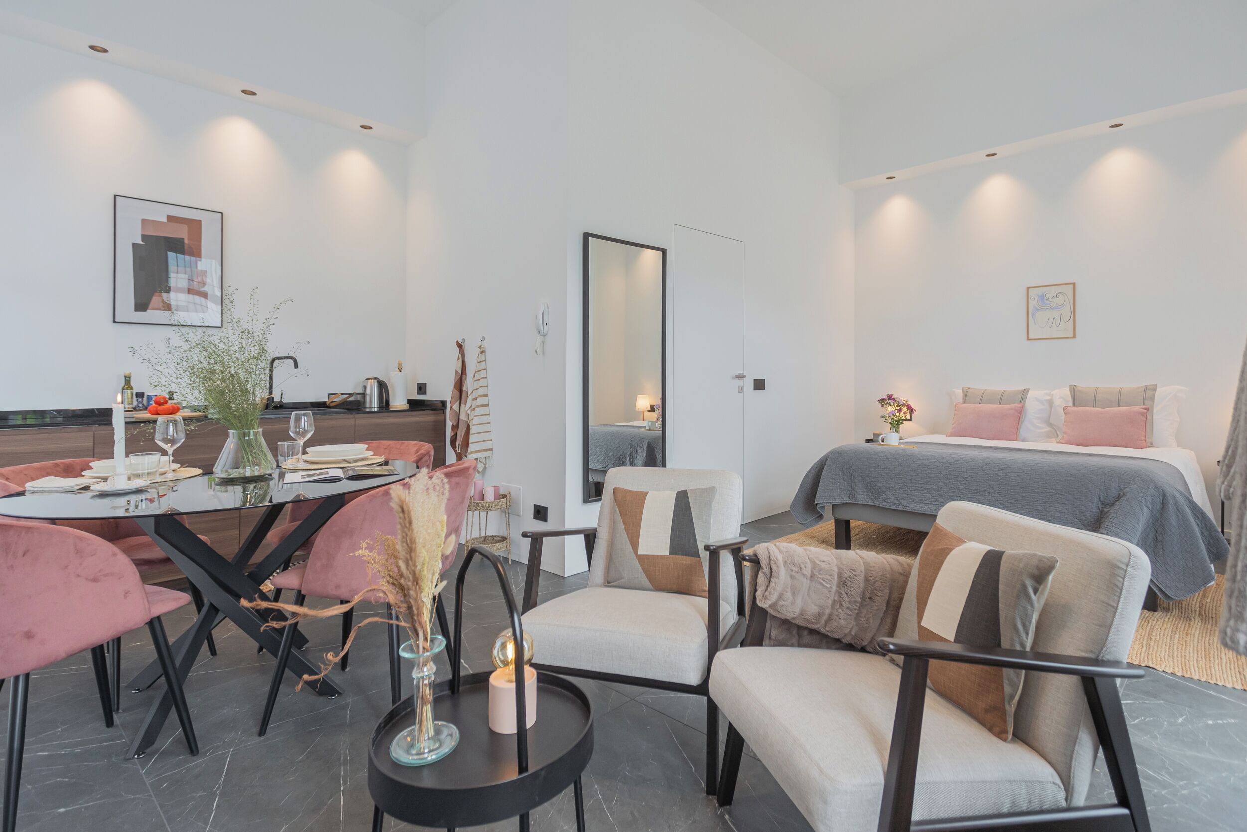 A modern, well-lit room with a dining table set for four, a sofa, and a bed with a gray and white bedding.