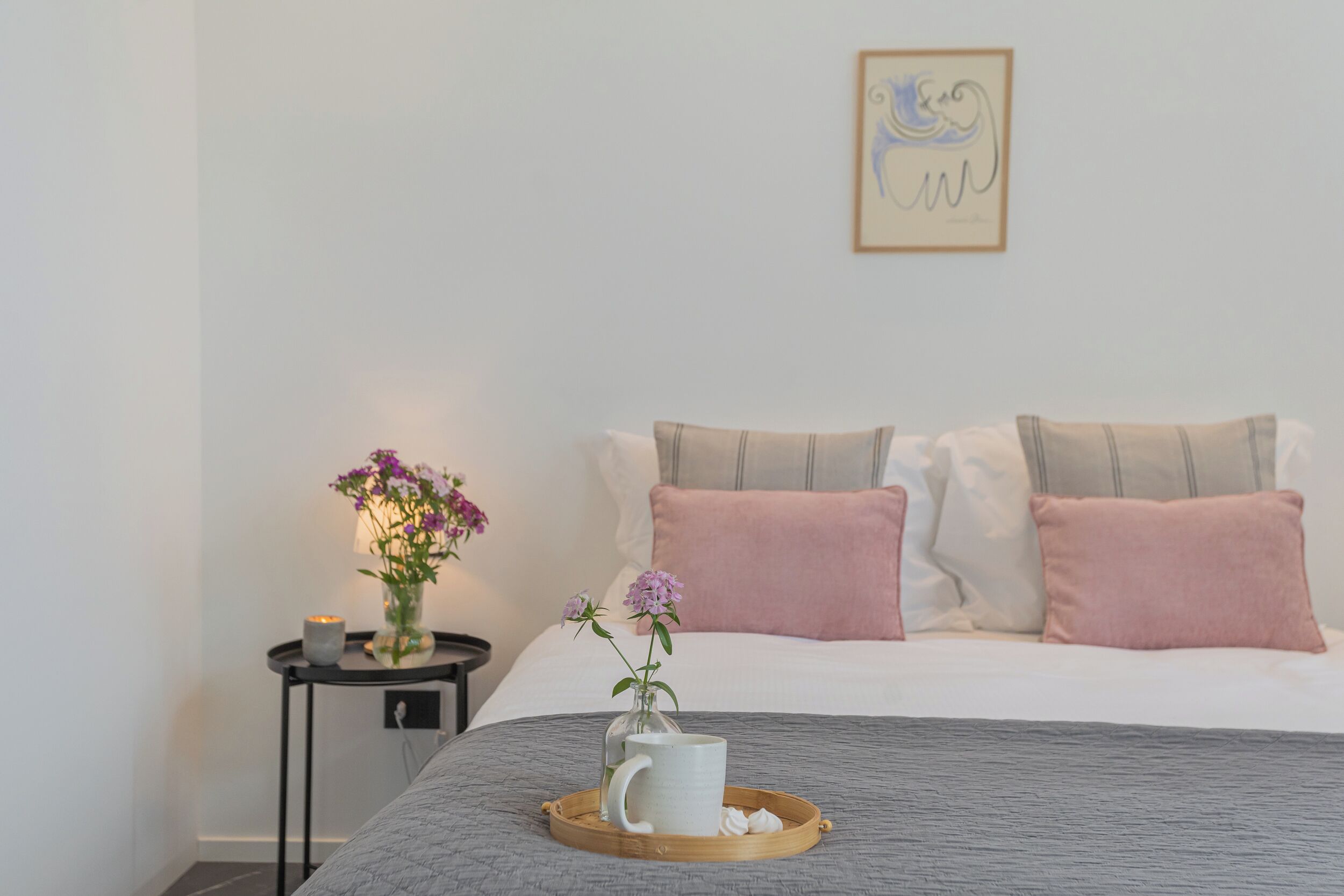 A neatly arranged bedroom with a bed, pillows, a bedside table with a vase of flowers, and a tray with a mug.