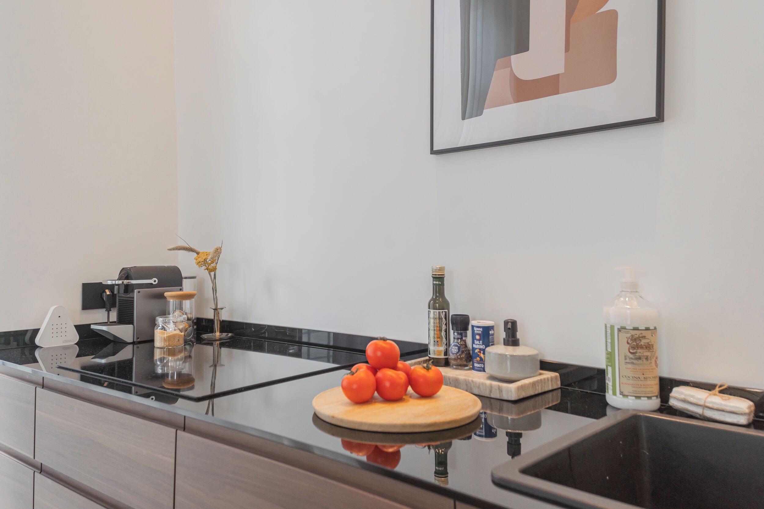 Una cocina moderna con encimera negra, tabla de cortar de madera con tomates, una cafetera y diversos utensilios de cocina.