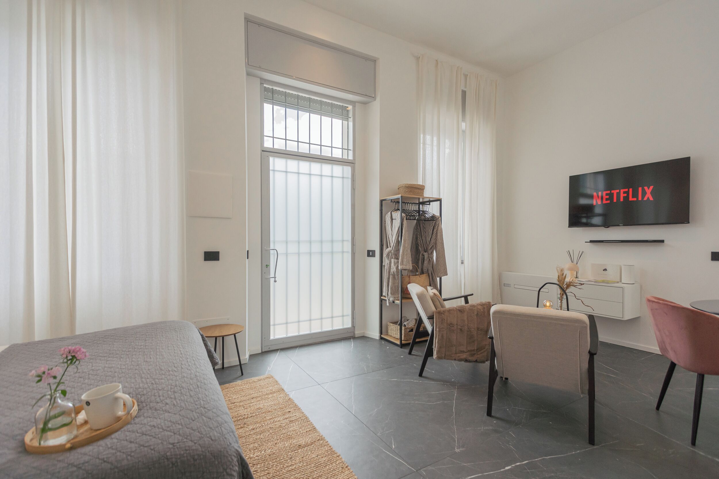 A modern hotel room with a bed, a TV displaying 'NETFLIX', a seating area with chairs, and a coat rack.