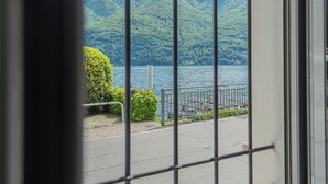 A view through a metal gate of a lake with a mountain backdrop, a paved walkway, and a railing. - Piccola Como Lotus by Rent all Como (Carate Urio)