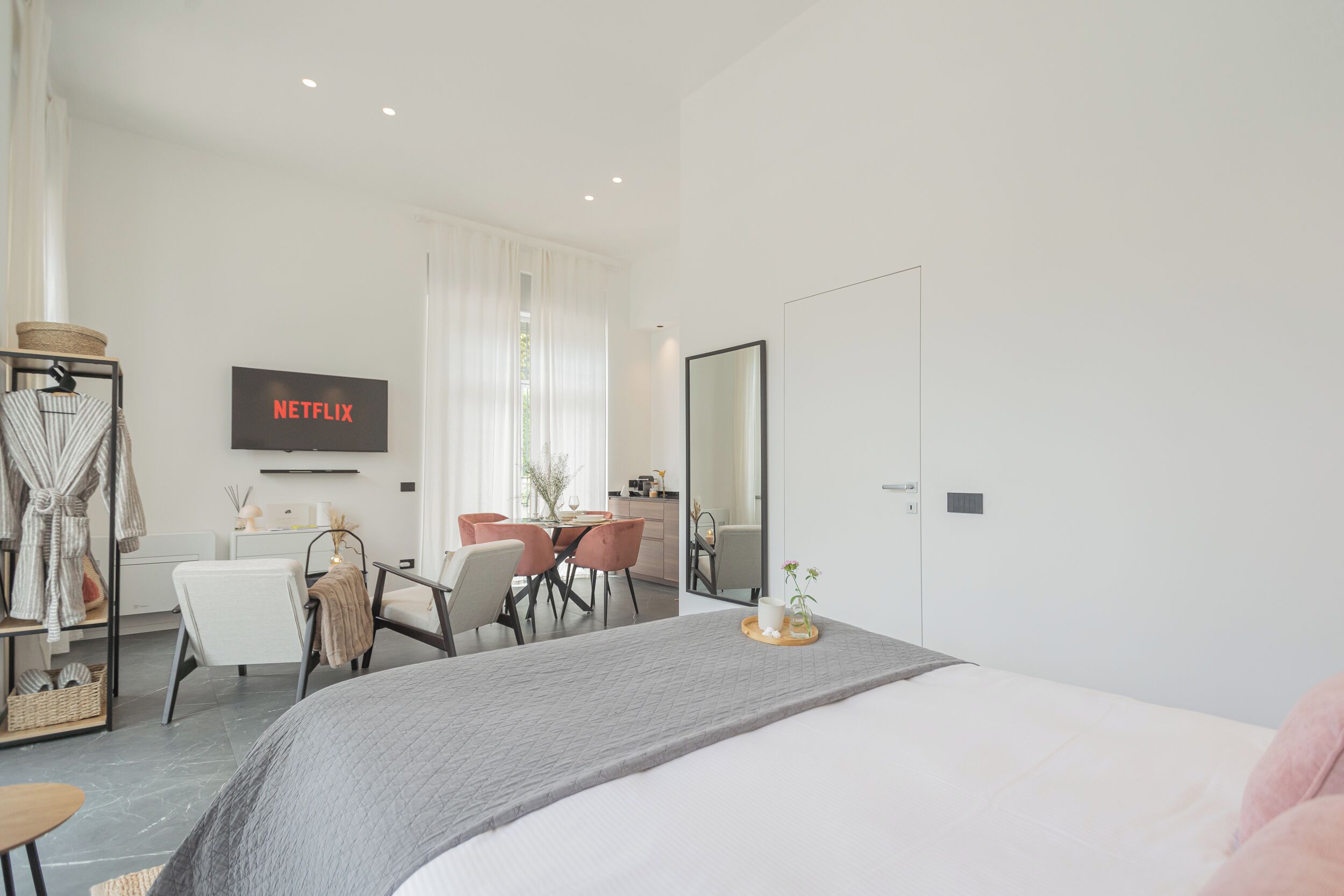 A modern hotel room with a large bed, a dining area with a table and chairs, a TV mounted on the wall, and a large mirror.