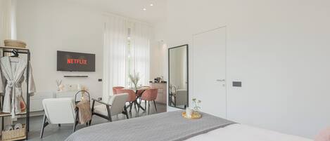 A modern hotel room with a large bed, a dining area with a table and chairs, a TV mounted on the wall, and a large mirror.