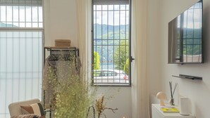 A modern room with a large window offering a view of a car and mountains, a flat-screen TV mounted on the wall, and a small table with a candle and decorative items. - Piccola Como Lotus by Rent all Como (Carate Urio)