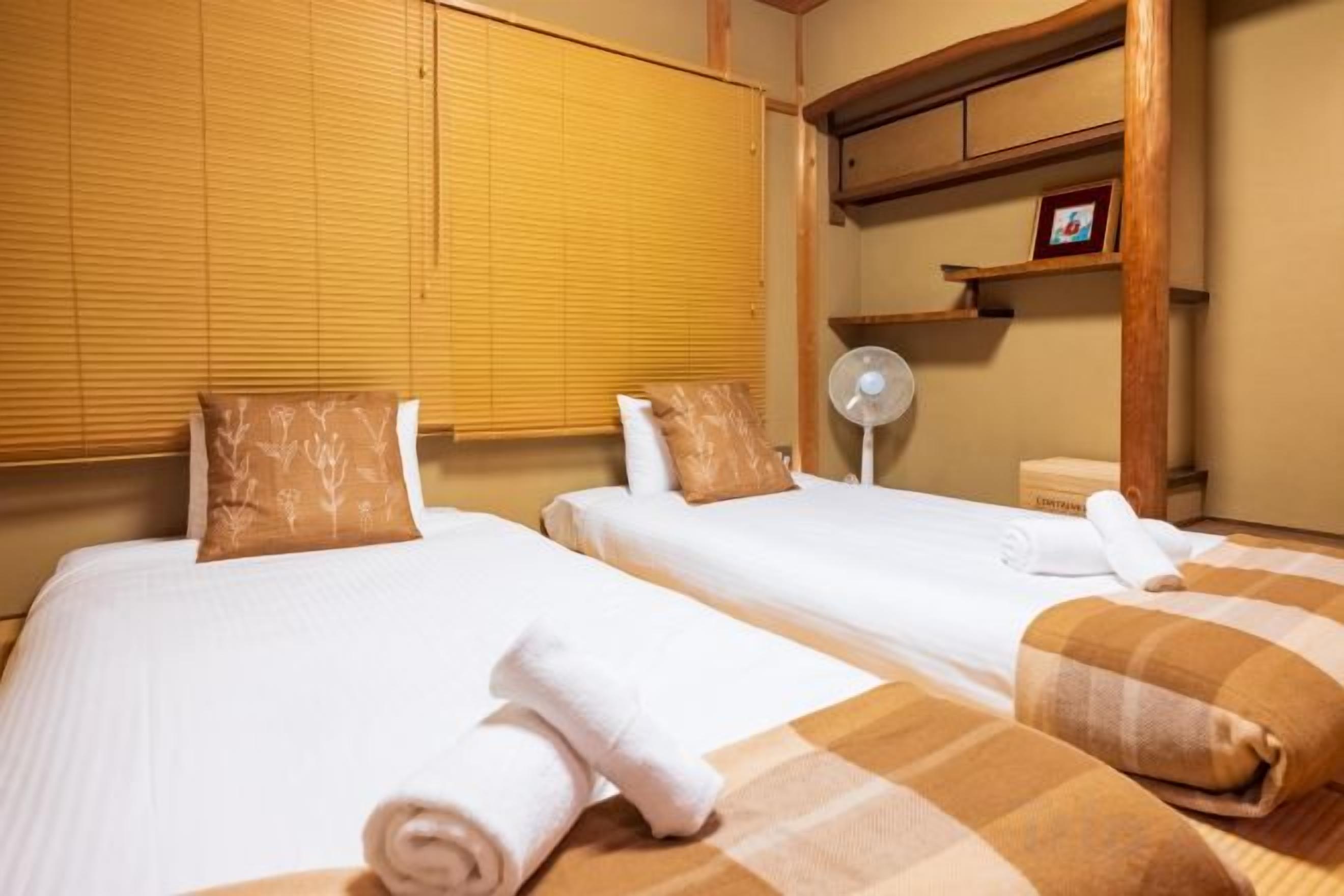 A hotel room with two beds, each with a pillow and a rolled towel. There is a bedside table and a small shelf with a picture frame.