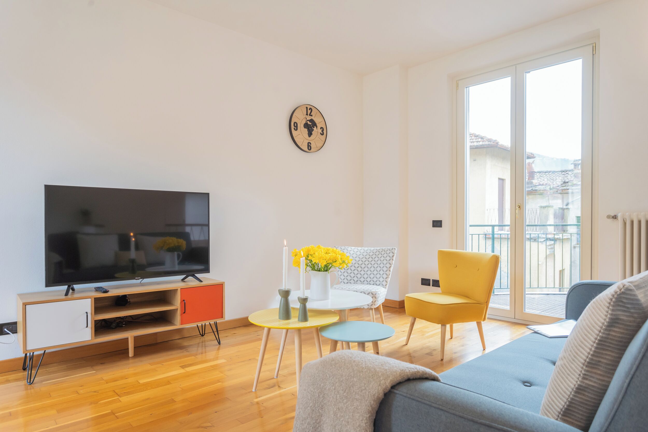 A modern living room with a flat-screen TV, a sofa, a yellow chair, and a small table with flowers.
