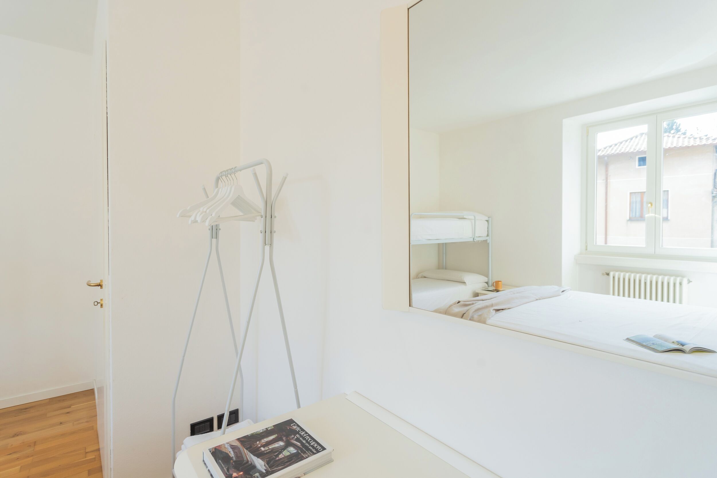 Une chambre avec un grand miroir, une table de chevet sur laquelle se trouve un livre, une fenêtre donnant sur des immeubles et un radiateur.