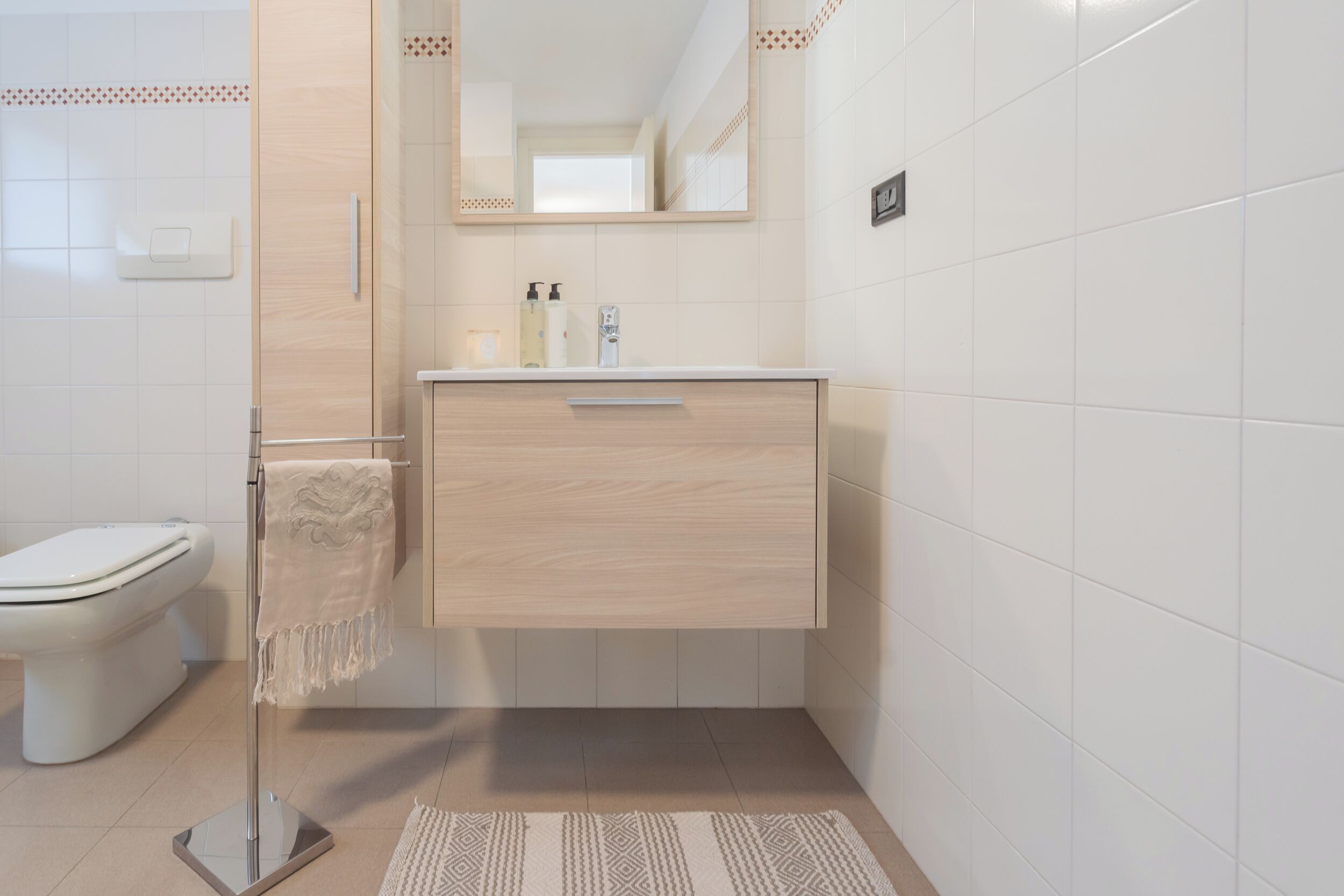 A modern bathroom with a white toilet, a wooden vanity with a mirror, and a patterned rug on the floor.