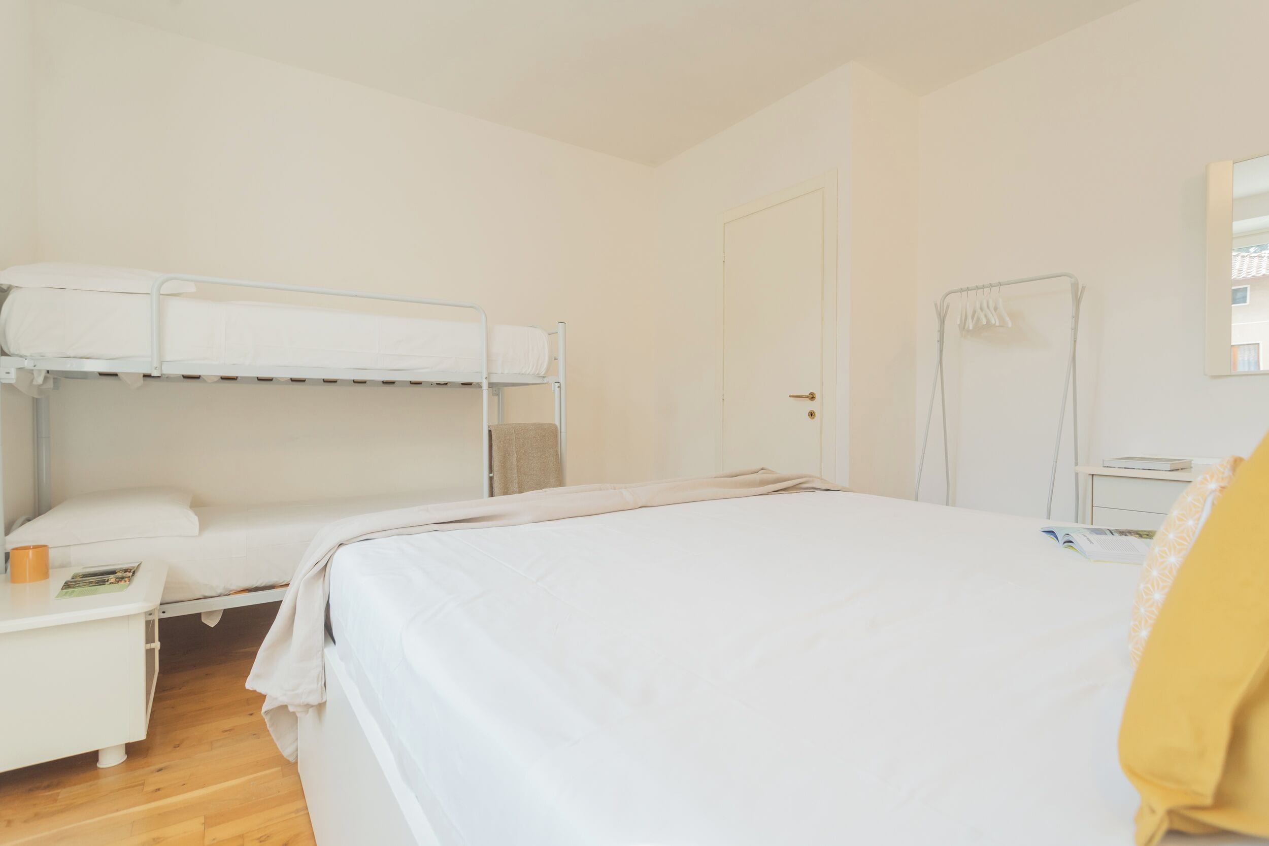 A room with a bunk bed, a single bed, a bedside table with a book and a mug, a towel rack, and a door.